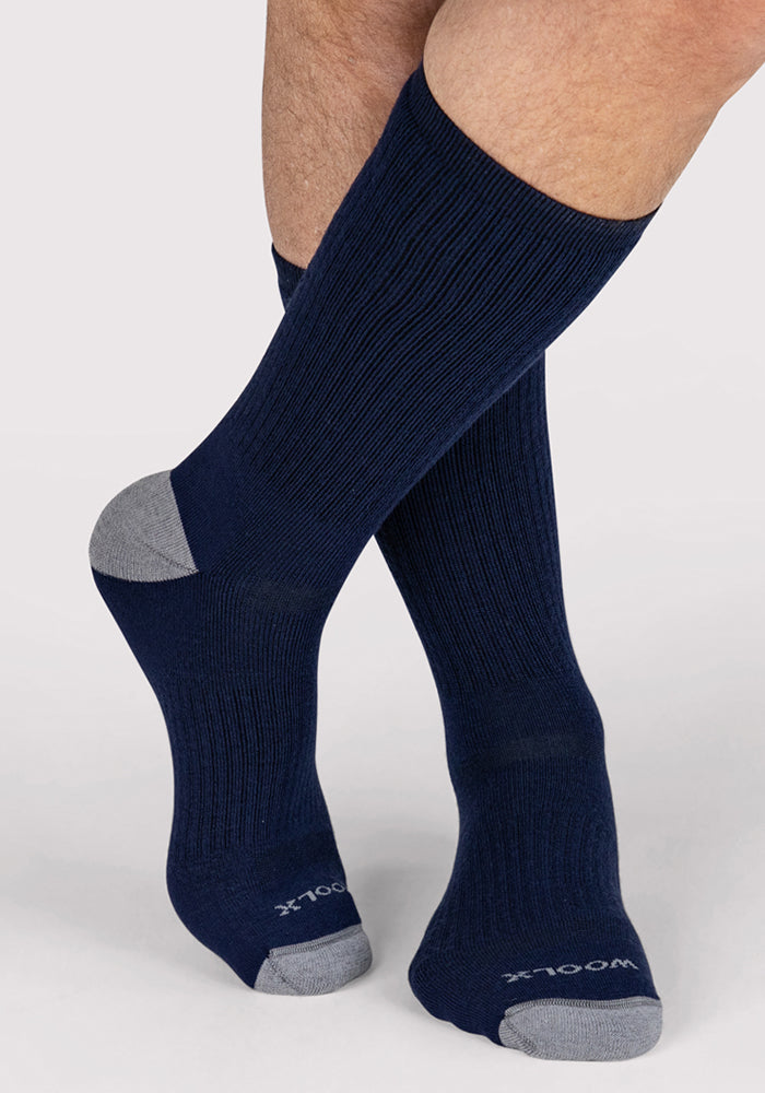 A pair of feet wears Woolx Mens Core Crew Socks in Deep Navy with gray heels and toes. These merino wool hiking socks have a ribbed texture, moisture-wicking properties, and "WOOLX" branding on the toes against a plain light gray background.