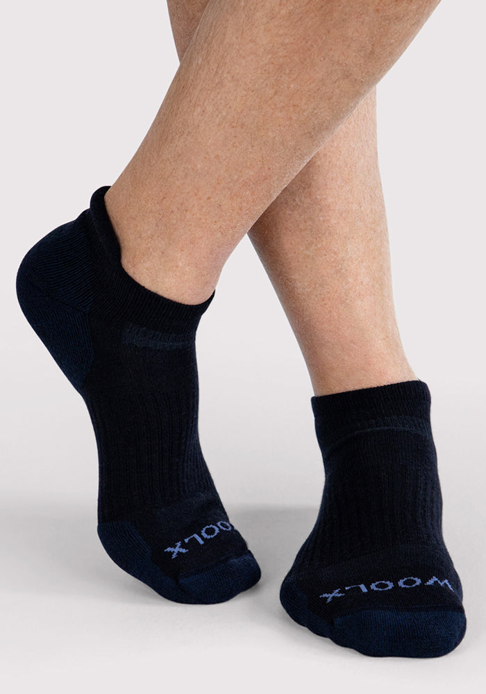A person with crossed legs wears Woolx Mens Rowan No Show Socks in Deep Night. These black, ribbed cushioned ankle socks feature "100X" text near the toes and are shown on a light gray background.