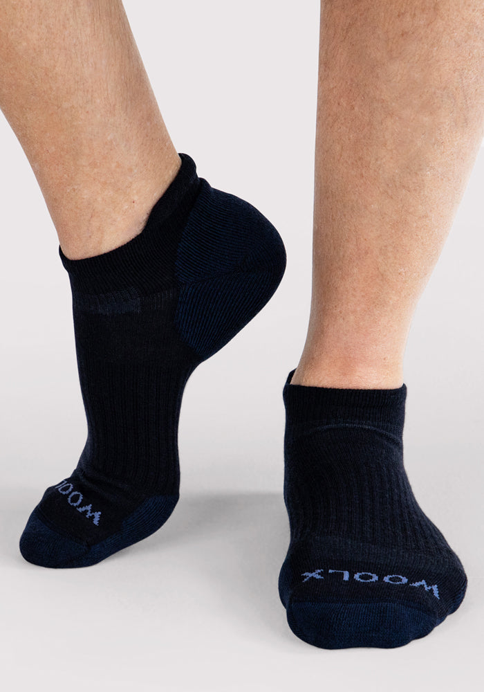 A person stands on tiptoe in Woolx Mens Rowan No Show Socks in Deep Night, with the brand name "Woolx" visible on the toes against a plain, light-colored background.