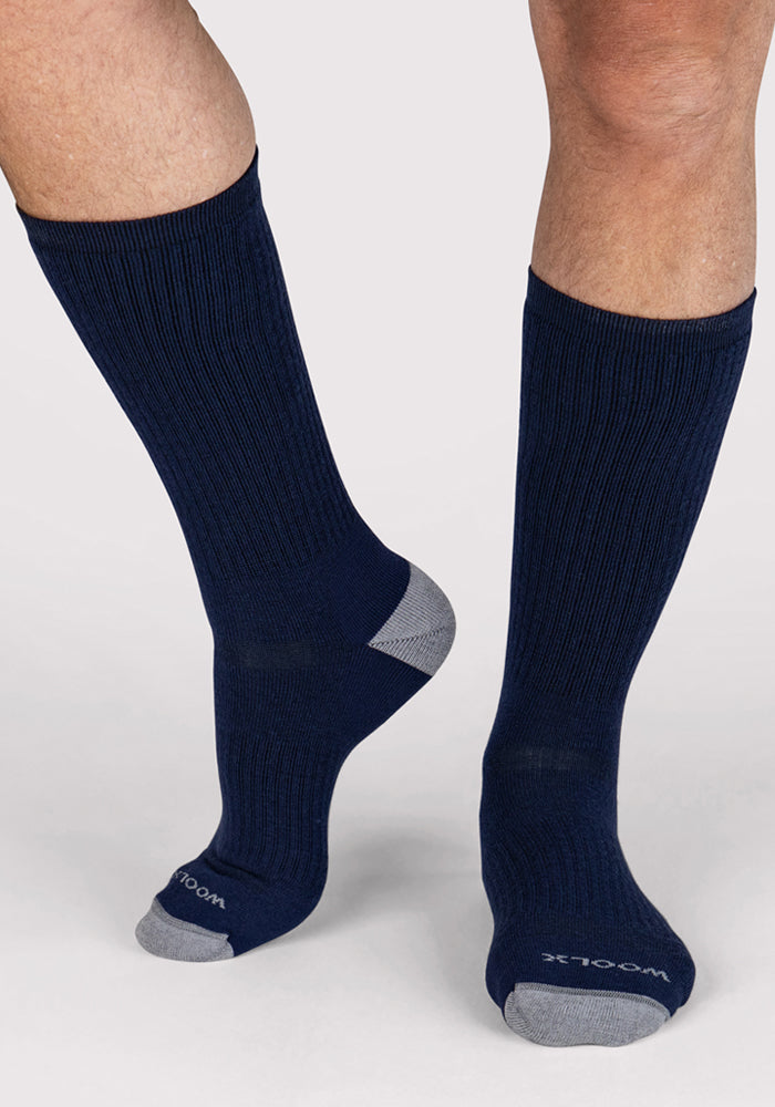 A person stands on a light background wearing Woolx Mens Core Crew Socks in Deep Navy, featuring gray toes and heels, "WOOLX" branding on the toe, and a moisture-wicking, performance fit.