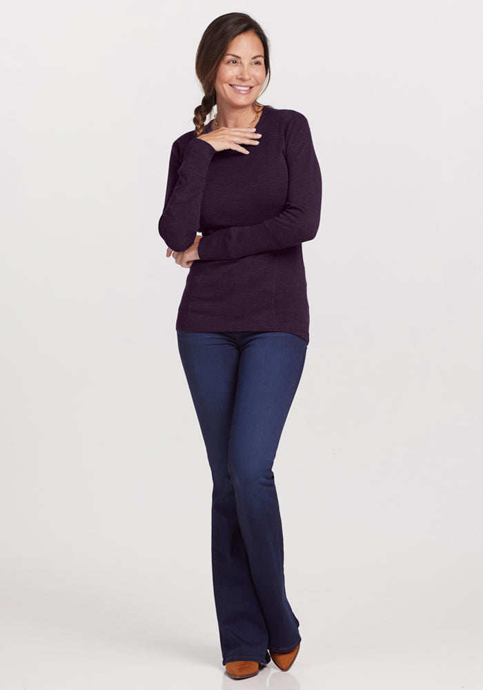 A woman with braided hair smiles with her hand near her chin, wearing the Woolx Hannah Long Sleeve Top in Deep Plum, dark blue jeans, and brown shoes, standing against a light background.