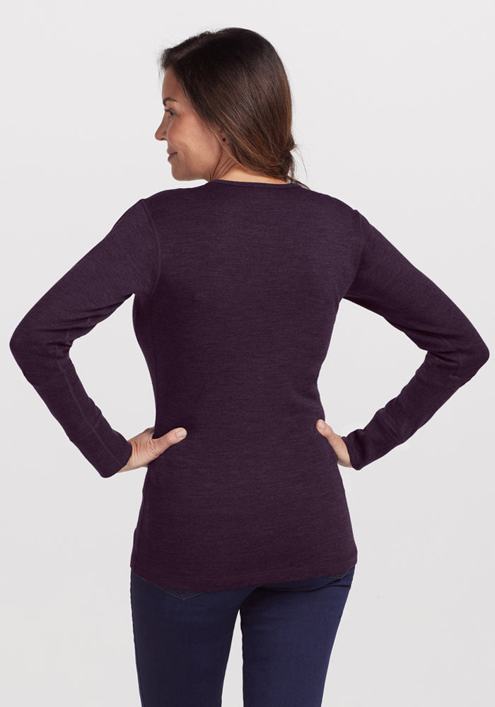 A woman with long brown hair, wearing the Woolx Hannah Long Sleeve Top in Deep Plum and dark jeans, stands facing away with her hands on her hips against a plain white background.