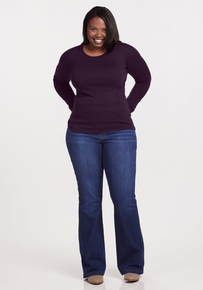 A woman stands smiling with her hands behind her back, wearing the Woolx Hannah Long Sleeve Top in Deep Plum, blue jeans, and beige shoes against a plain white background.