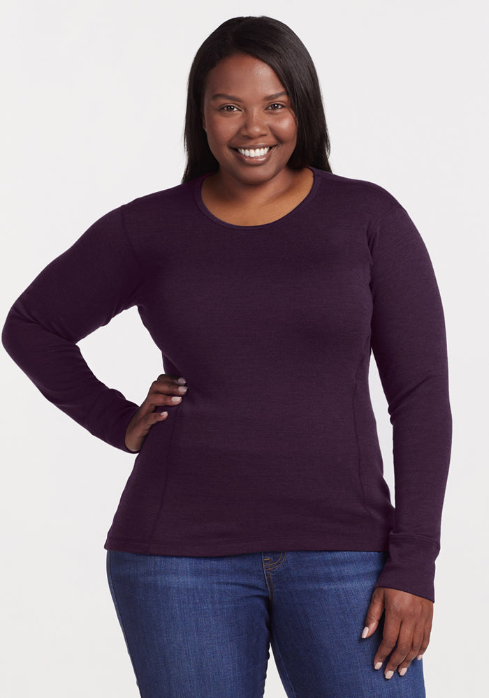 A woman smiles with one hand on her hip, wearing the Woolx Hannah Long Sleeve Top in Deep Plum and blue jeans against a plain white background. 
