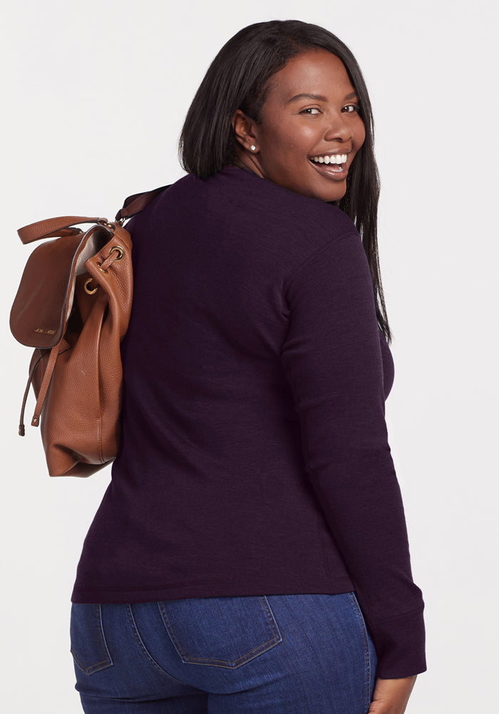 A smiling woman with long dark hair wears the Woolx Hannah Long Sleeve Top in Deep Plum and blue jeans, looking over her shoulder with a brown leather handbag against a plain white background.