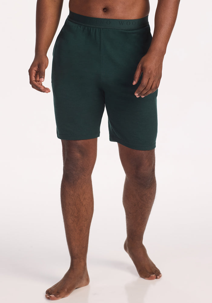 A person is shown barefoot, wearing Woolx Archer Sleep Shorts in Deep Timber and a matching Australian Merino Wool shirt, standing against a plain light background. The cropped image displays them from the shoulders down. 