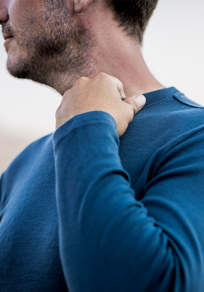 Wearing the Woolx Easton Crew Neck Shirt in blue, a person touches their neck with one hand, showing only the lower part of their face and upper torso against a blurred background.