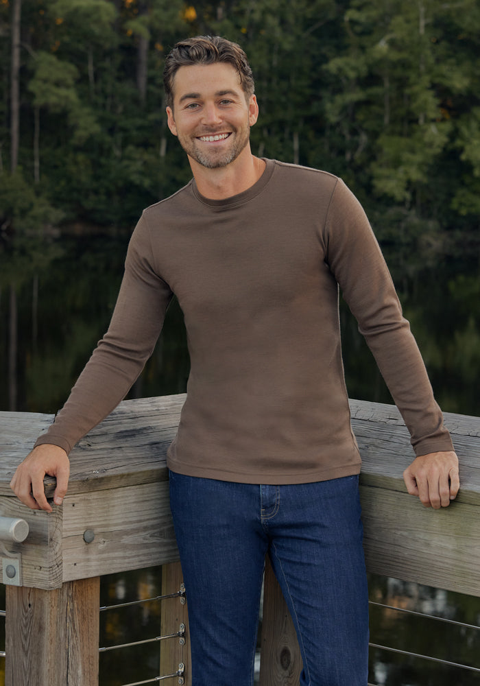 A man in the Woolx Easton Crew Neck Shirt - Bourbon Oak and blue jeans smiles as he leans on a wooden railing, with a lake and trees in the background.