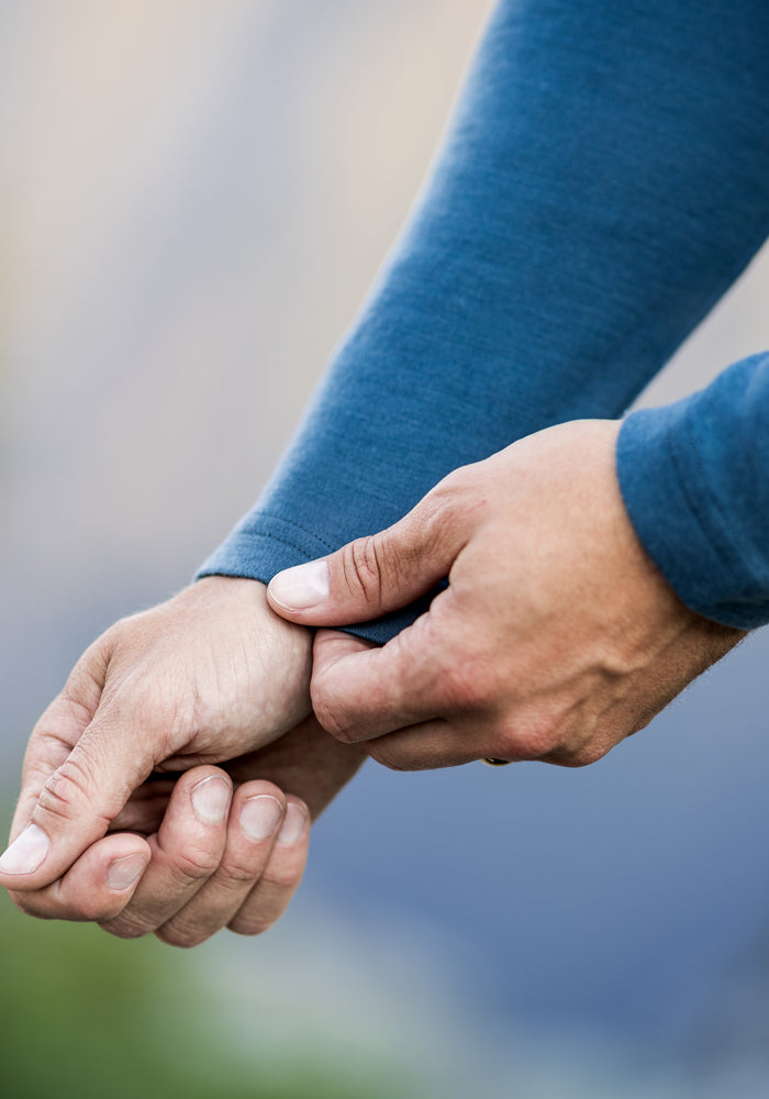 A close-up of a person wearing a blue Woolx Easton Crew Neck Shirt, pulling the sleeve down over their wrist with one hand against a blurred outdoor background.