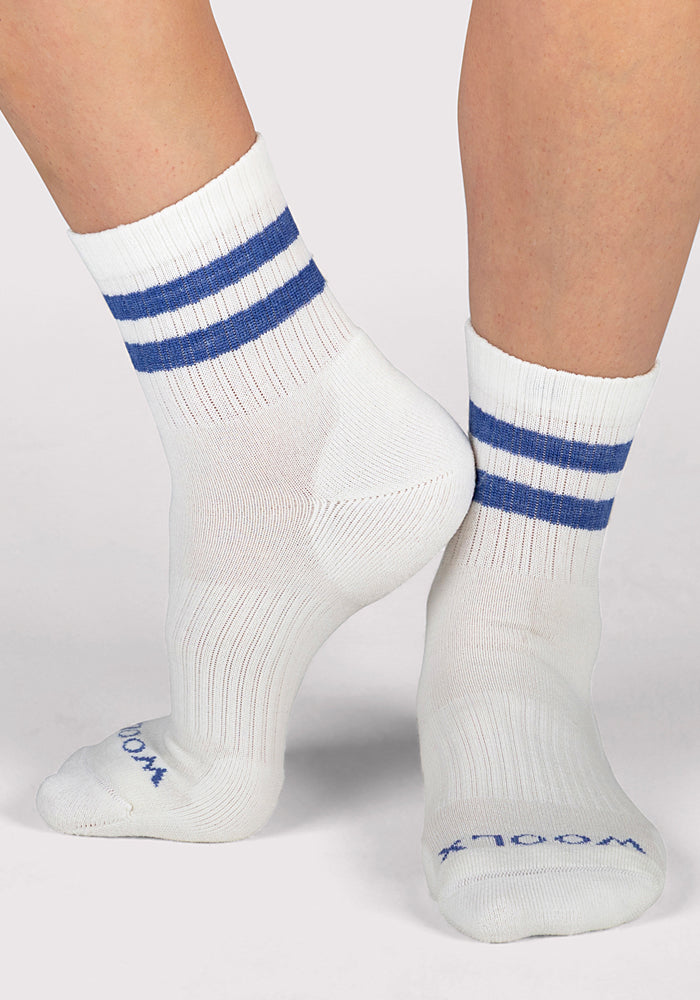 A person stands on tiptoe against a plain background, displaying the side and sole of Woolx Womens Varsity Mini Crew Socks in Electric Blue with two stripes and "WOOL" visible on the toe, showcasing their breathable design.