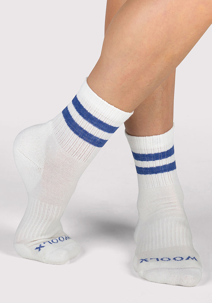 A person wears Womens Varsity Mini Crew Socks - Electric Blue by Woolx, featuring white merino wool with two blue stripes near the top and "WOOLX" in blue on the toes. Their legs are crossed against a plain light gray background.