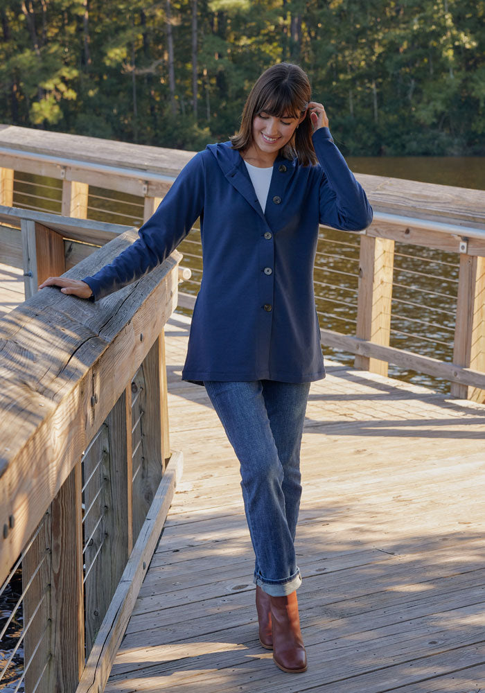 A woman wearing the Woolx Edna Hooded Cardigan in Deep Navy, with jeans and brown boots, smiles on a wooden dock by the water, touching her hair, with trees in the background.