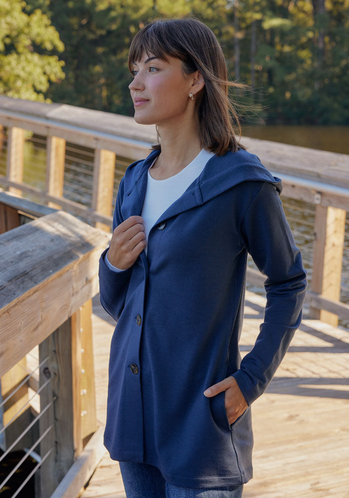 A woman stands on a boardwalk by the water, wearing the Woolx Edna Hooded Cardigan in Deep Navy over a white top. This women’s cold weather sweater keeps her warm as she gazes into the distance, trees behind her.