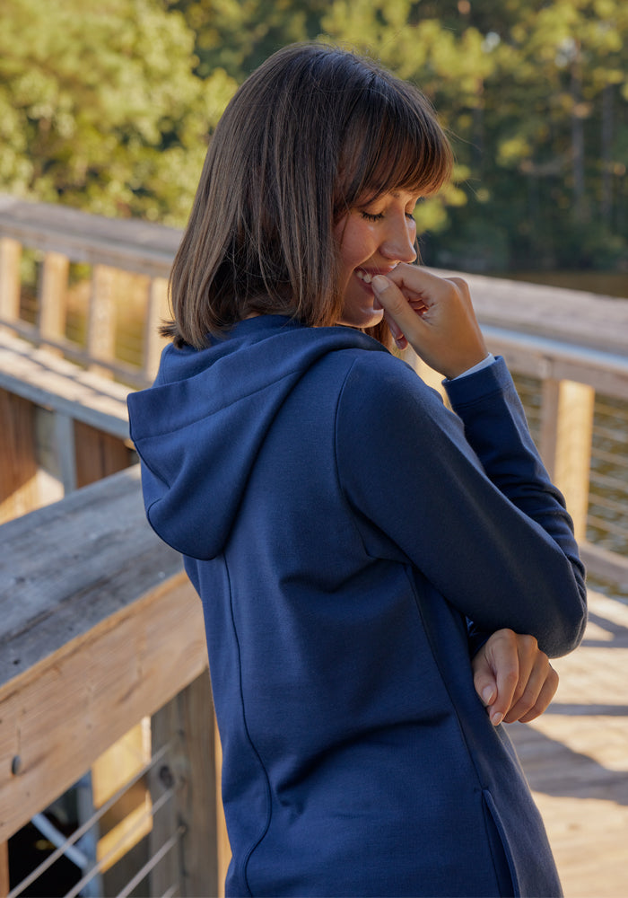 A woman wearing the Woolx Edna Hooded Cardigan in Deep Navy stands on a wooden boardwalk by the water, smiling with her hand near her mouth. Sunlight and trees brighten the background.