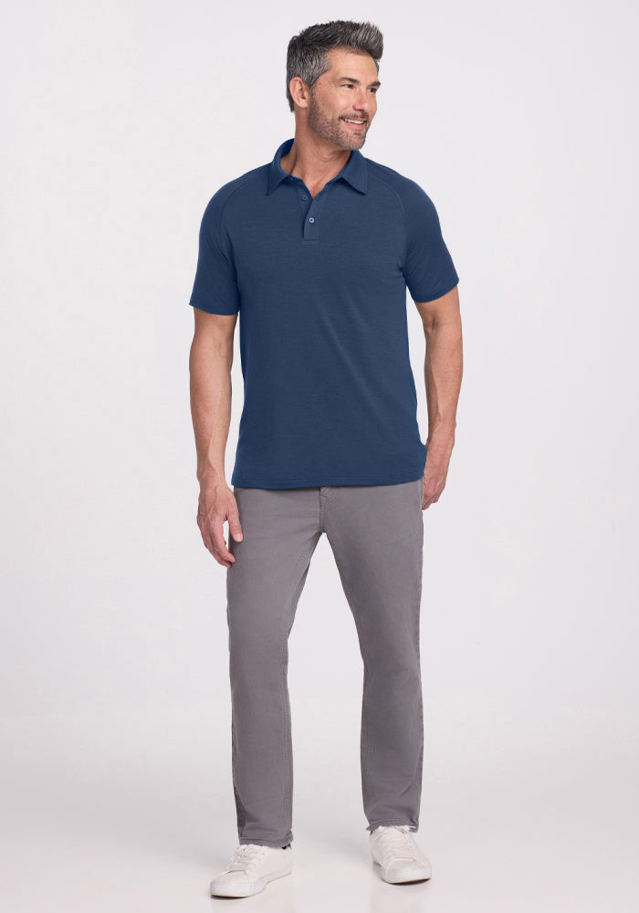 A man faces slightly sideways, smiling, in a Woolx Summit Polo - Starry Night (navy Merino wool with great moisture management), light gray pants, and white sneakers. He has short dark hair with gray streaks and stands before a plain light background.