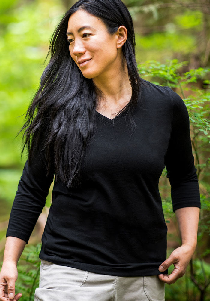 A woman with long black hair, wearing the Woolx Elena 3/4 Sleeve V Neck in black and light-colored pants, smiles outdoors while looking to the side in a lush green forest.