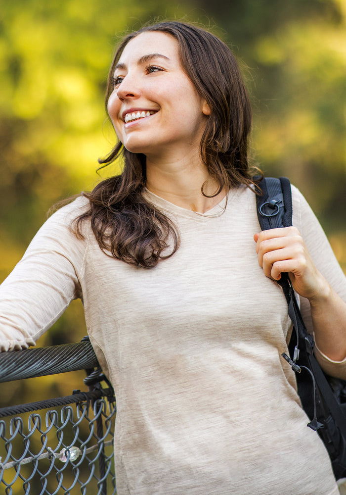 A woman with long brown hair smiles outdoors wearing the Woolx Elena 3/4 Sleeve V Neck in Cream Heather. She holds a black backpack strap and leans on a metal fence, with blurred greenery behind her.