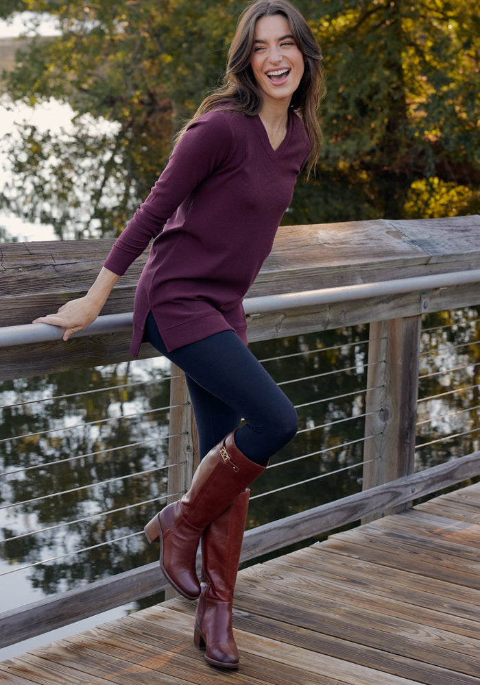 A woman smiles on a wooden bridge, wearing a maroon sweater, tall brown boots, and Woolx Ellis Seamless Tights in black, with trees and water in the background.