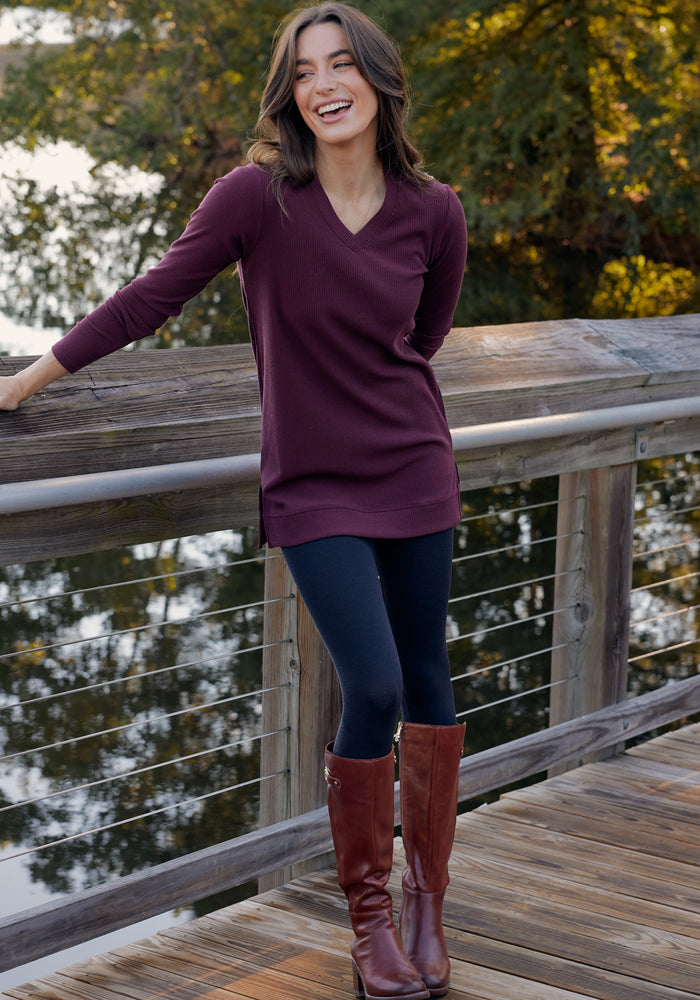 A woman smiles on a wooden bridge over water, wearing a maroon long-sleeve top, tall brown boots, and Woolx Ellis Seamless Petite Tights in black. Lush greenery and trees create a scenic background.