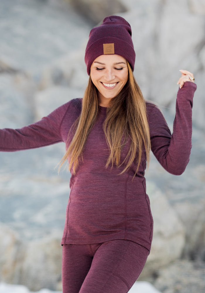 A woman wearing the Woolx Elsa Long Sleeve in Grape Harvest Melange and a matching beanie and leggings smiles with her eyes closed, standing outdoors among rocks and snow.