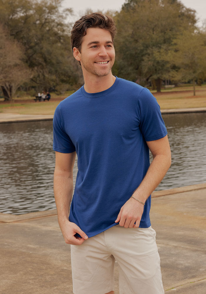A young man wearing the Woolx Endurance Tee in Estate Blue and beige shorts stands by a pond, smiling and looking to the side. Cloudy skies, trees, and park benches can be seen in the background.