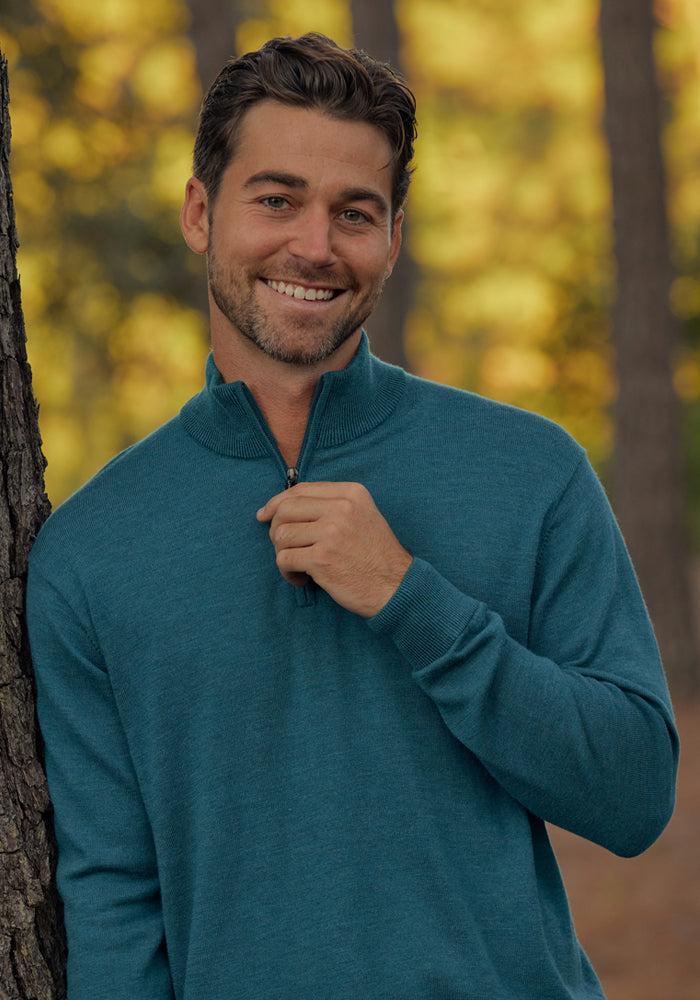 A man with short brown hair and a beard smiles outdoors among trees, wearing the Woolx Enzo Quarter Zip in Harbor Teal and gently holding the zipper collar. Soft, blurred autumn colors fill the background.