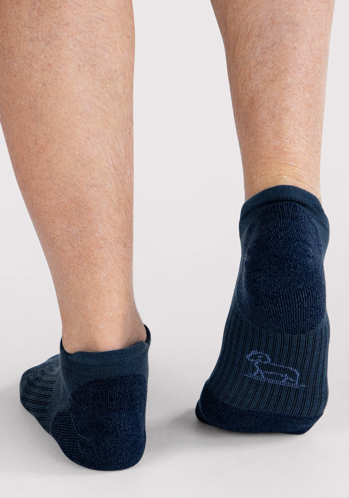 The image shows the back view of a person wearing Woolx Mens Rowan No Show Socks in Evening Tide, standing on a light surface. A small dog outline is visible on one of these cushioned Merino wool socks.