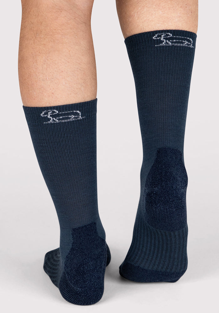 A close-up shows two legs in Woolx Mens Harbor Crew Sock - Evening Tide, navy blue moisture-wicking socks with a small white embroidered logo near the top, set against a plain light background.