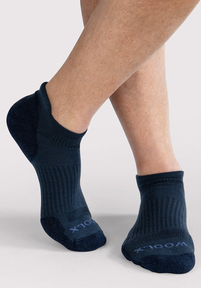 A person with light skin is wearing Woolx Mens Rowan No Show Socks in Evening Tide, featuring ribbed cuffs and contrasting textured toe and heel, while standing on a light gray surface with one foot crossed in front of the other.