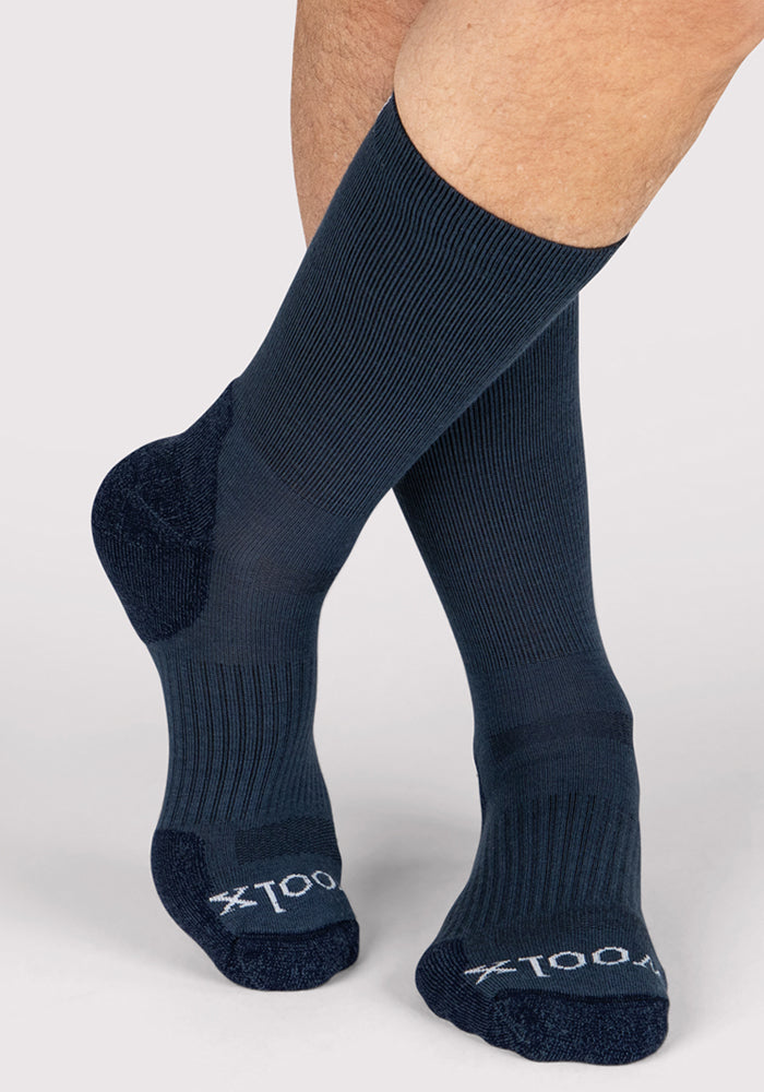A person wearing Woolx Mens Harbor Crew Socks in Evening Tide stands with feet crossed. "100X" appears on the toes of these odor-resistant, moisture-wicking socks featuring reinforced heels and toes.