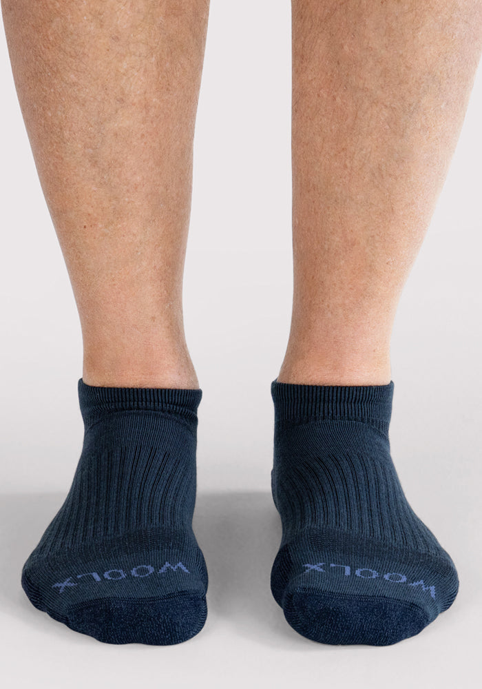 A pair of feet wearing Woolx Mens Rowan No Show Socks in Evening Tide, featuring light "WOOLX" lettering near the toes, standing on a light-colored surface.