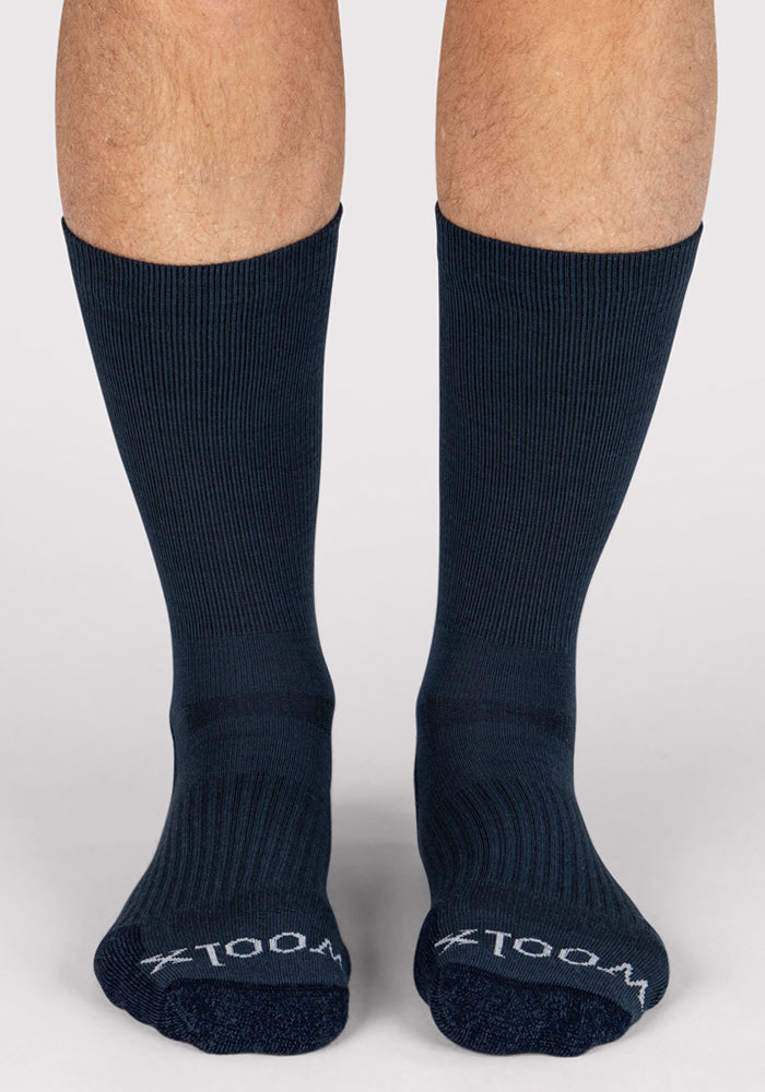 A pair of feet in Woolx Mens Harbor Crew Socks - Evening Tide, dark blue ribbed Merino wool with "WOOL" in white on the toes. These moisture-wicking socks are shown against a light gray background.