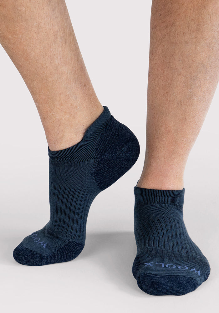 A close-up of a person wearing Woolx Mens Rowan No Show Socks in Evening Tide, with one foot slightly raised. The ribbed, moisture-wicking socks display "WOOL" on the toes against a plain light background.
