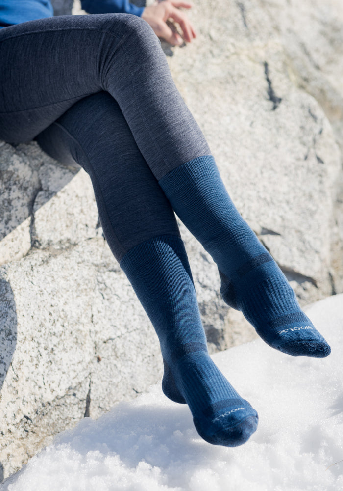 A person wearing Woolx Womens Harbor Crew Socks in black and gray leggings sits cross-legged on a rock, with snow visible on the ground nearby.