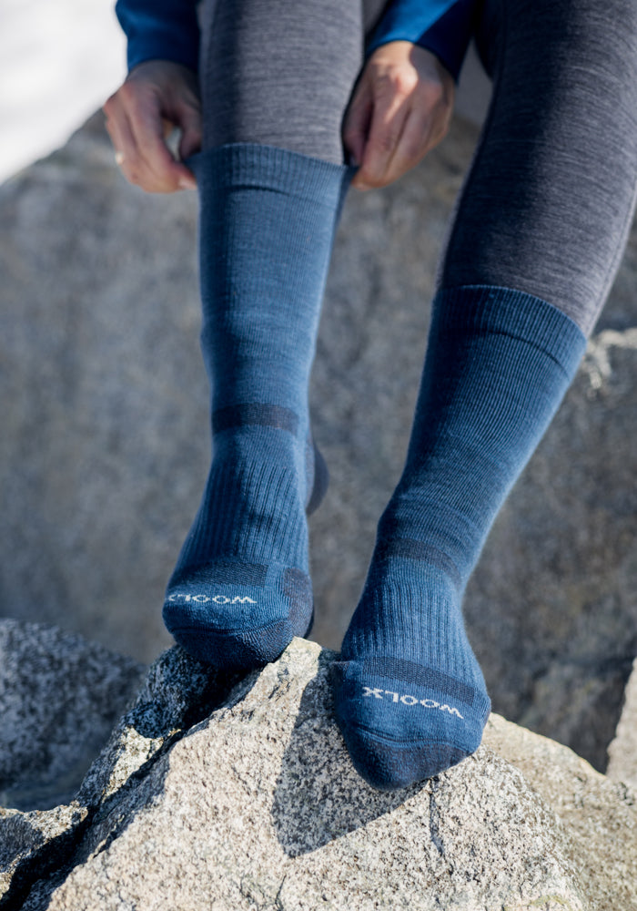 Outdoors on the rocks, a person pulls on Women's Harbor Crew Socks in Forest by Woolx over dark leggings, with "WOOLX" visible on the toes.