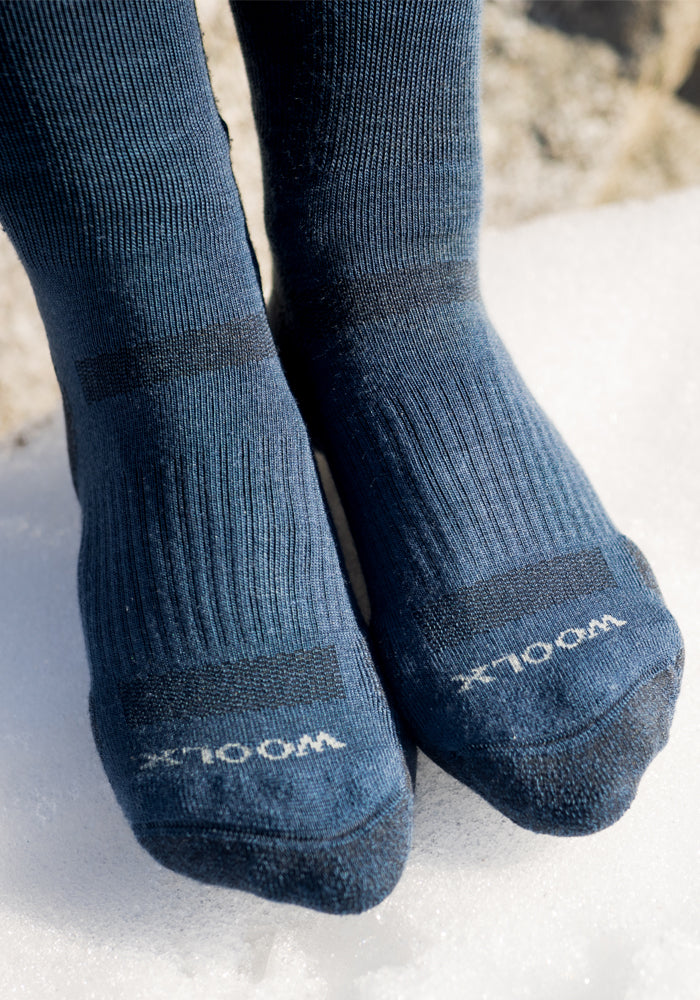 Close-up of feet in black Woolx Womens Harbor Crew Socks standing on snow. "WOOL" appears in white near the toes, showcasing their moisture-wicking and temperature-regulating features.