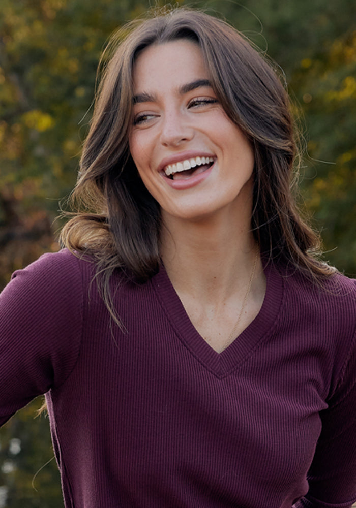 A woman with long brown hair smiles and looks to the side outdoors, wearing the Woolx Evie Ribbed Tunic in Majestic Purple. Trees are softly blurred in the background.