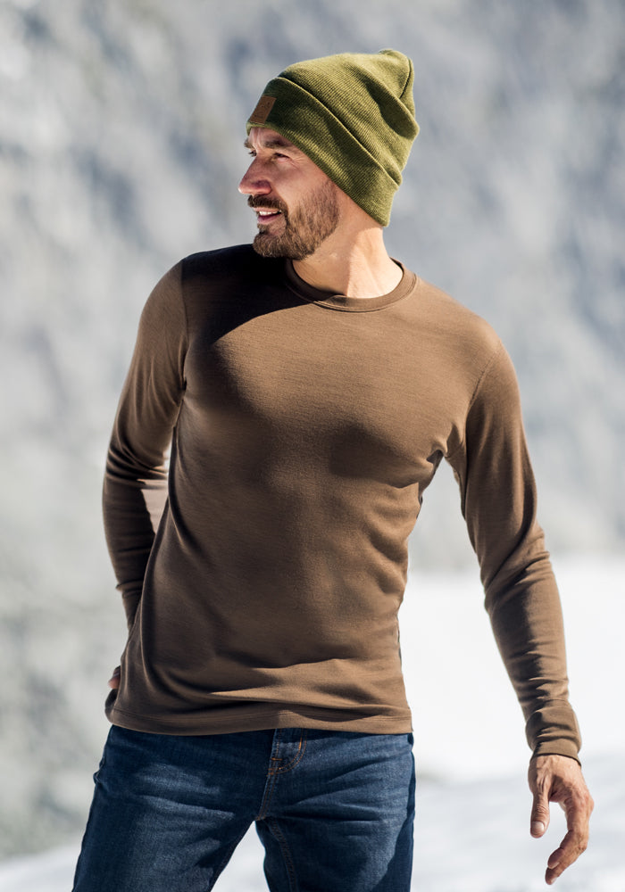 A man in a green knit beanie, Woolx Explorer Base Layer - Bourbon Oak, and blue jeans stands outdoors in a snowy, mountainous setting, looking to his left and smiling.