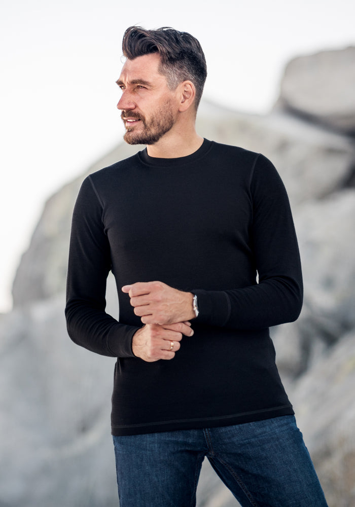 A man with dark hair and a beard, wearing the Woolx Explorer Base Layer in black and jeans, stands outdoors by light-colored rocks, looking to his right with a slight smile.