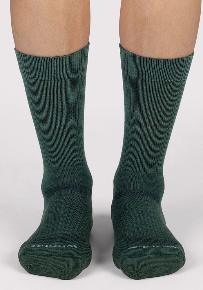A pair of feet, shown from the calves down, wear Woolx Womens Harbor Crew Sock in Forest—a tall, dark green Merino wool style—while standing on a white surface.