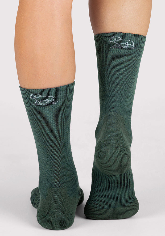 A close-up rear view shows two legs in Woolx Womens Harbor Crew Socks in Forest, featuring moisture-wicking fabric and a small white embroidered logo near the top, standing on a light gray surface.