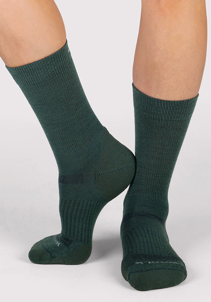 A close-up of a person wearing Woolx Womens Harbor Crew Sock in Forest, standing with one foot flat and the other heel slightly raised against a light gray background.