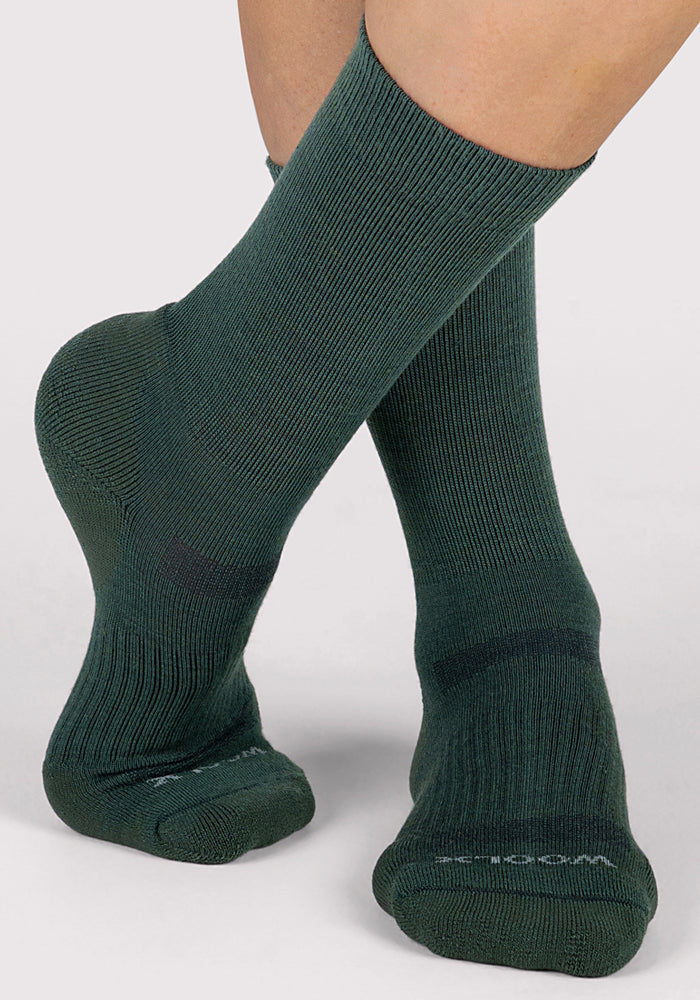 A close-up of two feet wearing Woolx Womens Harbor Crew Sock in Forest, ribbed and breathable, reaching mid-calf, with one foot slightly raised and crossed over the other against a light gray background.