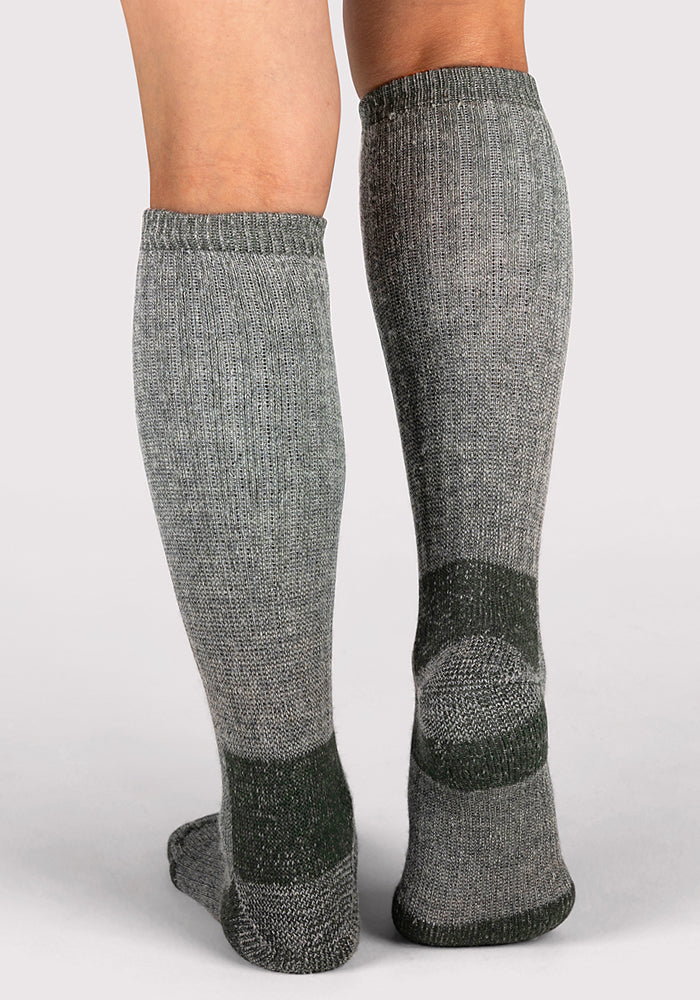 A pair of legs wearing Woolx Womens Extreme Over The Calf Socks in Forest Dark Moss, featuring dark gray accents at the heels, toes, and cuffs, shown from the back against a plain light background.