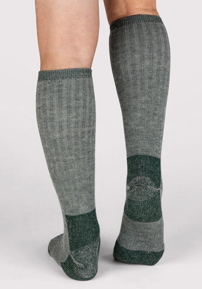 A person is wearing Woolx Mens Extreme Over The Calf Socks in Forest Dark Moss—gray knee-highs with dark green accents on the heels, toes, and cuffs—standing on a light surface with calves and feet visible.