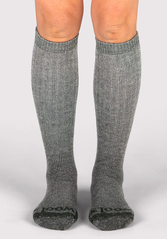 A pair of legs wears Woolx Womens Extreme Over The Calf Socks in Forest Dark Moss, standing on a neutral light background—ideal heavyweight merino wool socks for cozy cold weather comfort.