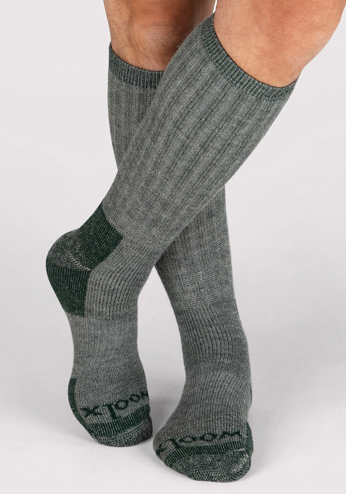 A person stands with legs crossed at the ankles on a light surface, wearing Woolx Mens Extreme Over The Calf Socks in Forest Dark Moss, featuring ribbed gray fabric and darker toe and heel accents.