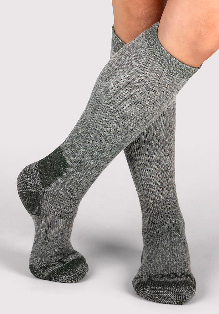 A person wearing Woolx Womens Extreme Over The Calf Socks in Forest Dark Moss, featuring darker gray accents on the toes and heels, stands against a plain light background.