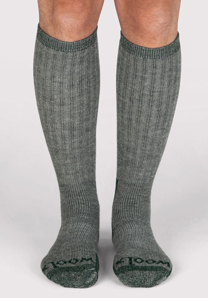 A pair of legs wearing Woolx Mens Extreme Over The Calf Socks in Forest Dark Moss, heavyweight merino wool, ribbed and knee-high, stand on a plain light-colored surface with feet facing forward.