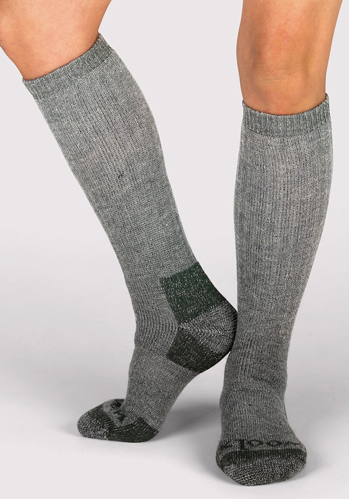 A person wears Woolx Womens Extreme Over The Calf Socks in Forest Dark Moss, featuring darker grey patches on the heel and toe, with one foot slightly lifted and crossed in front of the other against a plain background.